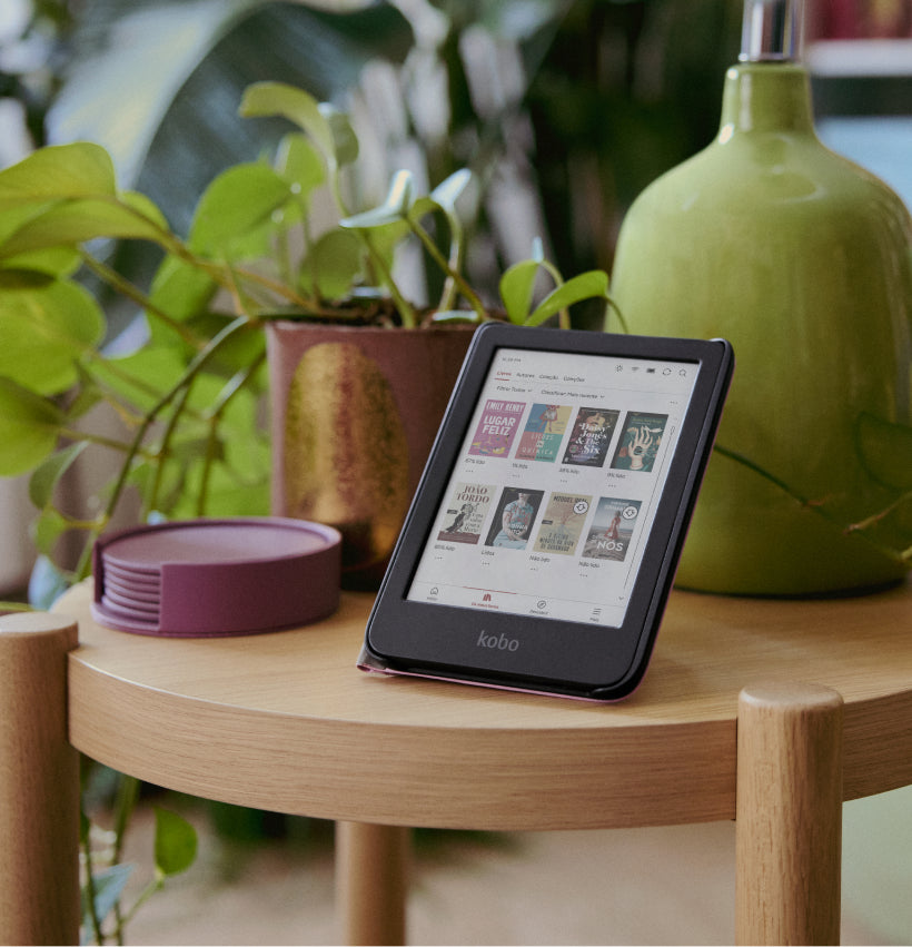 Um eReader Kobo Clara Colour no suporte da sua SleepCover, pousado numa mesa de madeira junto de um vaso com uma planta e um candeeiro.