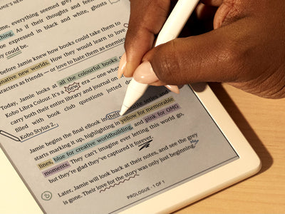 Uma mão é mostrada a usar a Kobo Stylus 2 para escrever diretamente num eBook num Kobo Libra Colour eReader numa superfície plana. Na página, vemos anotações e destaques coloridos.