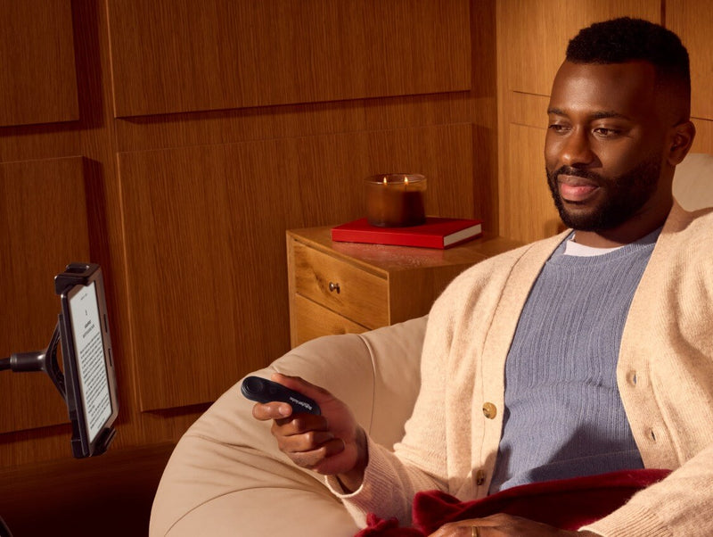 Um homem, confortavelmente sentado, segura o Kobo Remote na mão direita, enquanto olha para um eReader Kobo num suporte flexível.