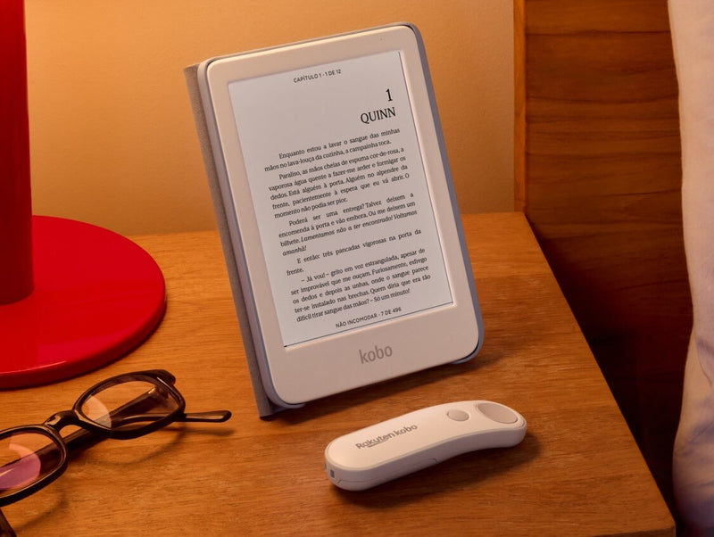 Um Kobo Clara Colour branco está apoiado numa mesa na sua SleepCover com suporte incorporado, ao lado de um Kobo Remote branco e um par de óculos.