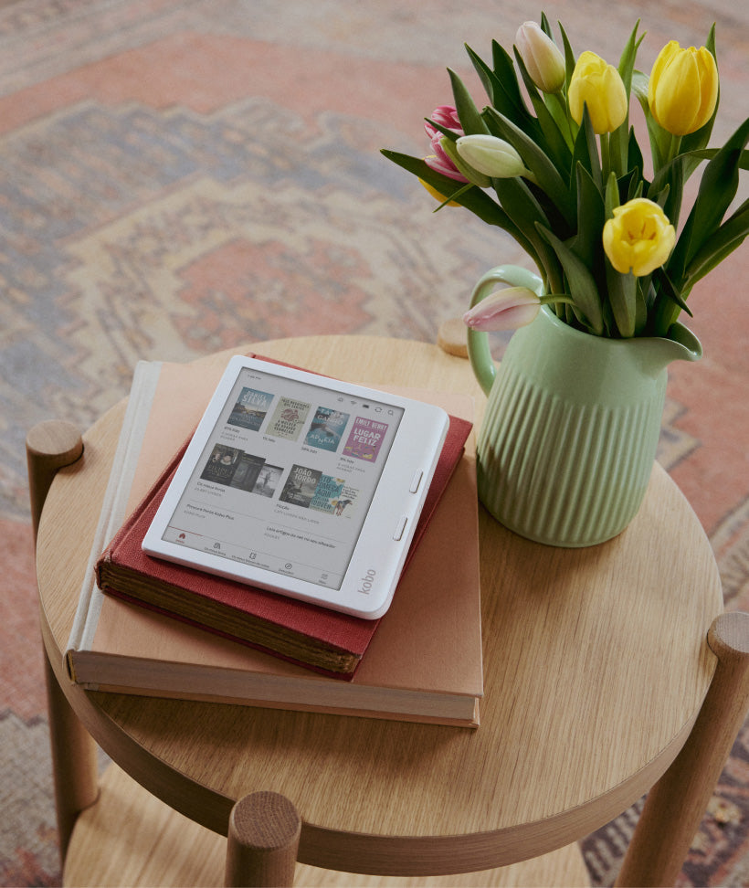 Um eReader Kobo Libra Colour pousado sobre dois livros de capa dura, numa mesa de madeira ao lado de flores numa jarra verde.